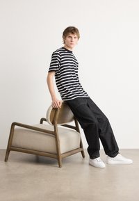 Tricou striped alb-negru, blugi negri din denim, adidași albi; sprijinit de un scaun din lemn cu suport din material textil deschis. Fundal neutru.