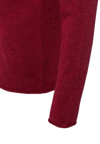 Nahaufnahme eines roten Strickpullover-Ärmels und des unteren Torsobereichs, die das strukturierte Gewebe und die Seitennahtdetails zeigen.