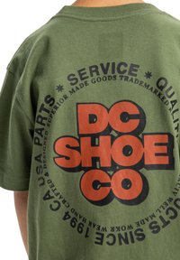 Olivově zelené bavlněné tričko s velkým černým a oranžovým grafickým prvkem "DC SHOE CO", obklopené kulatým textem.