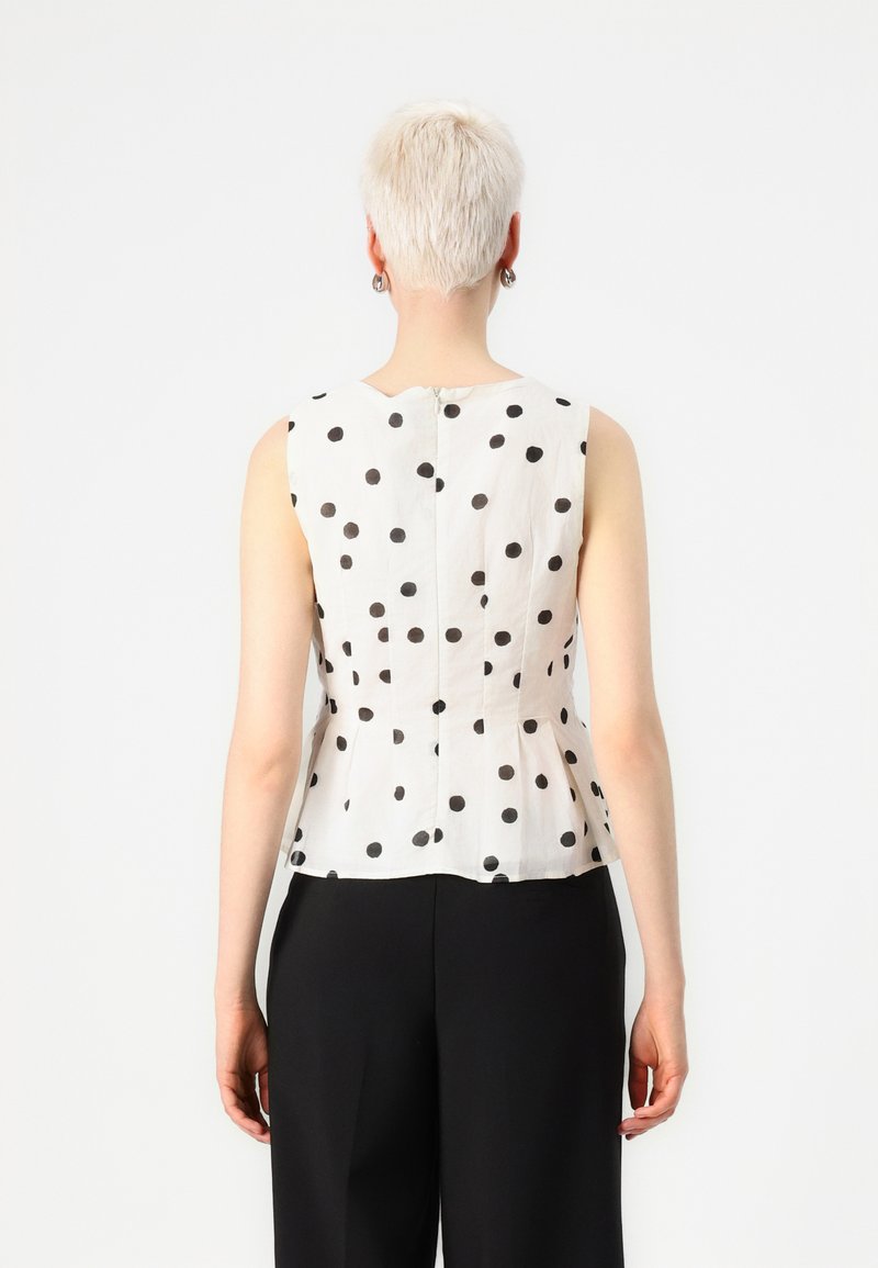 Top sans manches en tissu blanc avec des pois noirs, présentant un buste ajusté, un ourlet peplum et une fermeture éclair au dos.
