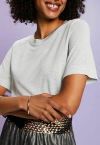 Personne souriante portant une chemise gris clair scintillante à manches courtes, une ceinture tissée métallique et un bracelet argenté, les bras croisés devant un fond violet clair.