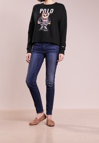 Černá mikina s grafickým designem medvěda, modré úzké džíny a hnědé mokasíny. Měkká tkanina, volný střih a elastické manžety na rukávech.