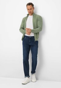 Green zip-up cardigan over a white T-shirt, paired with dark blue jeans and white sneakers. Smooth texture, fitted design.