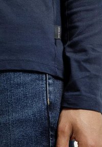 Granatowy t-shirt z długim rękawem o gładkiej fakturze i boczną metką; w połączeniu z dopasowanymi niebieskimi dżinsami o wyblakłym wzorze.