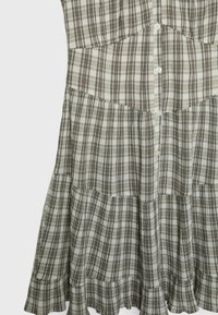 Koszulowa sukienka w przygaszonych odcieniach beżu i brązu. Posiada zapięcie na guziki, warstwowy dół z falbanami oraz miękką teksturę materiału.