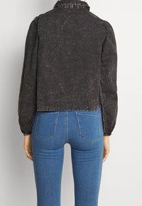 Czarna, krótka kurtka z bufkowymi rękawami, wykonana z fakturowanej tkaniny, z kołnierzem i surowym wykończeniem u dołu; zestawiona z niebieskimi, dopasowanymi jeansami z tylnymi kieszeniami.