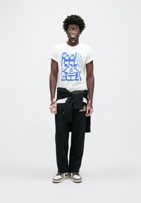 Biele tričko s modrým grafickým dizajnom, čierne tepláky a sivobiele tenisky. Sveter zviazaný okolo pása pridáva detail.