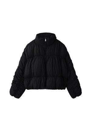 Jeune femme portant une doudoune noire, un crop top noir, un pantalon noir avec des bandes blanches sur les côtés et des chaussures noires, debout devant un fond rouge.