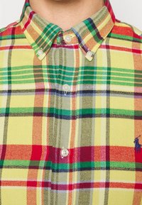 Károvaná košile na knoflíky ve žluté barvě, s červenými, zelenými a modrými pruhy. Vyrobena z látky s mírně texturovaným povrchem a viditelným logem polo.