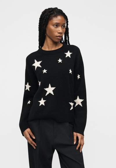 Donna con lunghi capelli intrecciati che indossa un maglione nero con motivi a stelle bianche e pantaloni neri, in piedi davanti a uno sfondo semplice.