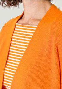 Pomarańczowy dziergany kardigan o fakturowanym wykończeniu, noszony na białej koszuli w pomarańczowe paski. Zwróć uwagę na otwarty przód i prążkowane zdobienia.