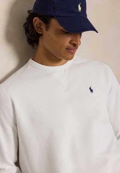 Ung mand iført en hvid sweatshirt og en mørkeblå kasket, begge med et lille polo-spiller logo, kigger ned med et let smil.