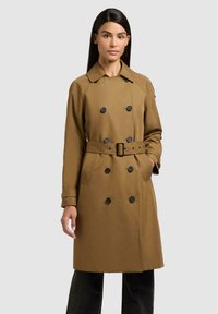 Donna che indossa un trench marrone doppiopetto con bottoni neri e una cintura coordinata, in piedi davanti a uno sfondo grigio chiaro uniforme.