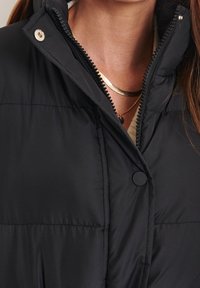 Svart dunjacka med hög krage, utrustad med dragkedja och tryckknappsstängning. Insidan är fodrad med ett mjukt material som har en strukturerad yta.