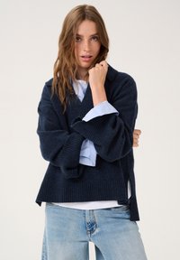 Pulover tricotat bleumarin cu decolteu în V, cu manșete cu dantelă și slituri laterale, purtat peste o cămașă albastru deschis, împreună cu blugi deschiși la culoare.