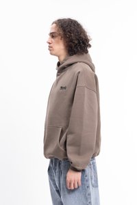 Barna kapucnis pulóver elülső zsebbel, hímzett logóval és bordázott mandzsettával. Világoskék farmernadrággal viselve. Háttér sima fehér.