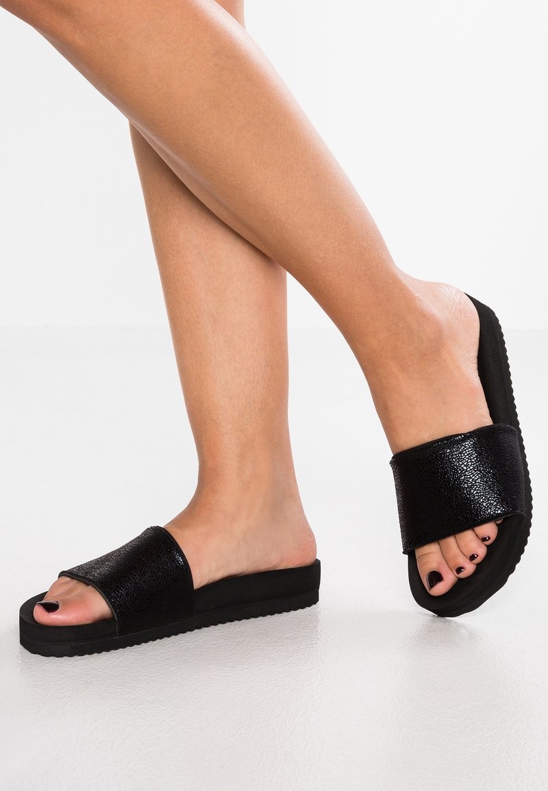 Jambes portant des sandales slides noires texturées avec des semelles épaisses, orteils peints en foncé, debout sur une surface blanche lisse.
