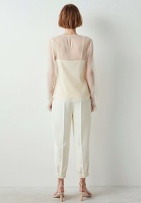 Przezroczysta, jasnobeżowa bluzka z teksturowanej tkaniny o prostym kroju, zestawiona z białymi zwężanymi spodniami z mankietem i dopasowaną talią.