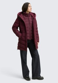 Bordeaux gewatteerde parka met een capuchon en een rits aan de voorkant, voorzien van een gewatteerd ontwerp en zijzakken, gecombineerd met donkere flared jeans en zwarte laarzen.