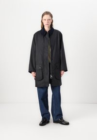 Casaco preto com colarinho, dois bolsos frontais e fecho com botão. Usado sobre uma blusa verde e combinado com calças largas e sapatos pretos.