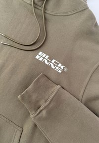 Beige hoodie with drawstrings; features "BLCK BNNS" logo in white. Soft fabric, ribbed cuffs, and hem. Accent stitching visible.