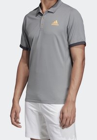 Šedé polo tričko s límcem, krátkými rukávy a oranžovým logem Adidas. Má tmavý lem na rukávech a zapínání na knoflíky.