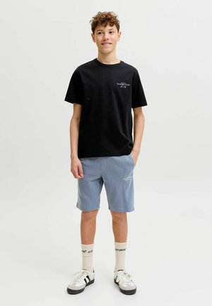 Garçon adolescent debout, portant un t-shirt noir, un short bleu clair, des chaussettes blanches avec texte, et des baskets blanches avec des rayures noires, sur un fond uni.