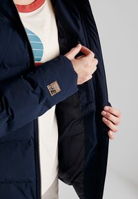 Tmavě modrá prošívaná bunda s hladkým povrchem, zapínáním na zip a štítkem na rukávu. Je vidět vnitřní kapsa, bunda je nošena přes světle barevnou košili.