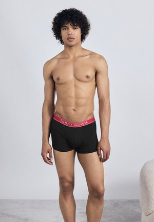 Jauns vīrietis ar cirtainiem matiem stāv kails, ģērbts melnās Tommy Hilfiger bokseršortos ar sarkano jostu minimālistiskā telpā.