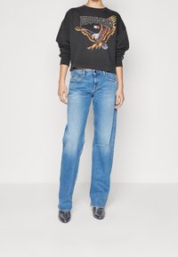 Zwarte sweatshirt met een adelaar graphic en tekst, gecombineerd met lichtblauwe, losse jeans met lichte slijtage. Zwarte puntige schoenen.