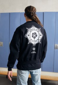 Hanorac negru cu un grafic alb mare pe spate, text într-un design circular și manșete ribbate. Purtați peste blugi de un albastru deschis.