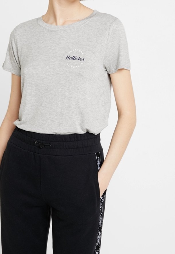 Šedé tričko s krátkým rukávem, na hrudi s malým modrým logem "Hollister", doplněné černými tepláky s bočními akcenty.