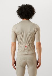 Maillot de cyclisme beige à manches courtes avec un tissu texturé, coupe ajustée, et une poche arrière avec un logo rond. Porté sur des legging assortis.