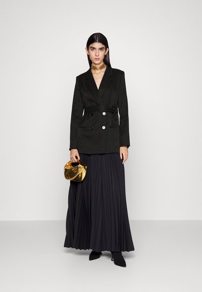 Zwarte dubbelgeknoopte blazer met een strik in de taille, gecombineerd met een zwarte geplooide rok en een glanzende gouden handtas. Zwarte puntige laarzen maken de look compleet.