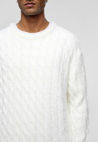 Weißer Kabelstrickpullover mit rundem Halsausschnitt, der eine weiche Textur und lange Ärmel aufweist. Das Material wirkt flauschig mit einem erhabenen Muster.