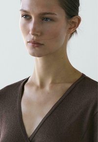 Brun V-hals top med ribbet tekstur. Modellens ansigt er delvist synligt med naturlig makeup og glat hår. Blød, neutral baggrund.