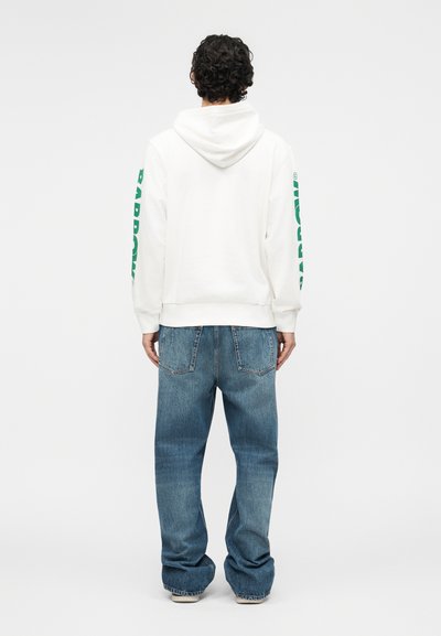 Άνδρας με σγουρά μαλλιά, που φοράει λευκή φούτερ με πράσινο κείμενο στα μανίκια και φαρδιά μπλε τζιν, στέκεται με την πλάτη σε φόντο μονόχρωμο.