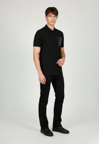 Czarna koszulka polo o gładkiej fakturze, z kieszenią o wyraźnej teksturze. W zestawie z czarnymi, dopasowanymi spodniami i czarnymi trampkami.