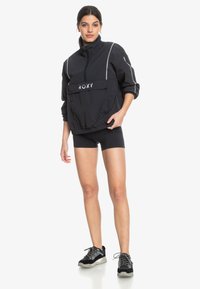 Sort vindjakke med høj krave, frontlomme og "ROXY" logo i hvid. Har reflekterende striber og elastiske manchetter. Sammen med sorte shorts og sneakers.