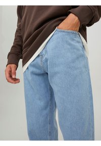 Jean en denim bleu clair avec une coupe décontractée, comportant une poche arrière, associé à un sweat-shirt marron foncé superposé sur une chemise blanche.