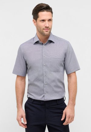 Uomo che indossa una camicia a maniche corte color grigio chiaro con taschino sul petto e pantaloni scuri, in piedi davanti a uno sfondo bianco uniforme.