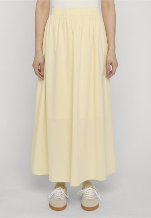 Person iført en lys gul, lang nederdel med elastisk talje, beige sneakers med hvide snørebånd og lysegule sokker, stående mod en ensfarvet baggrund.