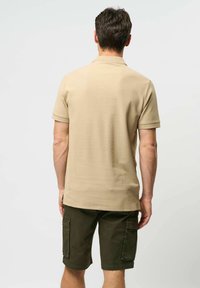 Muž otočený zády, oblečený v béžovém polo tričku s krátkým rukávem a tmavozelených cargo šortkách, na jednobarevném světlém pozadí.
