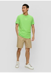 Grünes Baumwoll-T-Shirt mit kurzen Ärmeln und rundem Halsausschnitt, mit einem kleinen weißen Logo auf der Brust. Kombiniert mit beigen Shorts und weißen Turnschuhen.