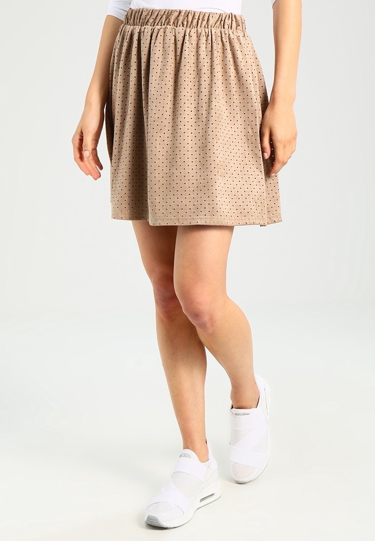 Falda beige con cintura elástica, con pequeños lunares marrones, fabricada en un tejido ligero y con forma acampanada. Combinada con zapatos blancos.