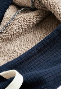 Tissu texturé bleu marine rencontre un intérieur en polaire beige doux, avec un design matelassé et un cordon de serrage de couleur claire contrastante.