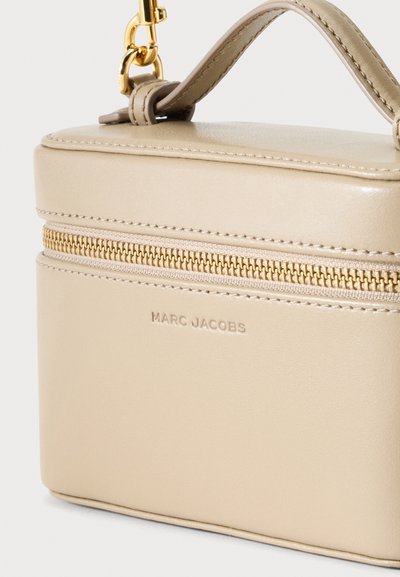 Μπεζ ορθογώνια τσάντα Marc Jacobs με χρυσό φερμουάρ, επάνω χερούλι και χρυσά μεταλλικά στοιχεία σε ουδέτερο φόντο.
