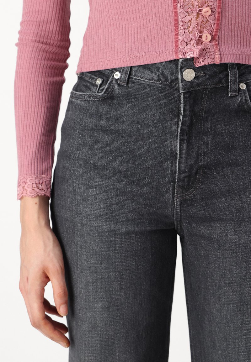 Haut rose à côtes avec des accents en dentelle au niveau des manches, associé à un jean en denim foncé. Le jean présente des détails en métal argenté et un design classique à cinq poches.