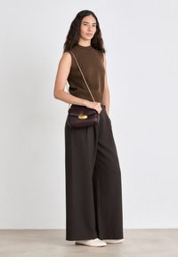 Top smanicato a maglia marrone abbinato a pantaloni larghi scuri. Una piccola borsa bordeaux con un accento dorato è indossata a tracolla. Calzature bianche.