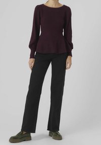 Frau in einem dunkelvioletten langärmeligen Peplum-Pullover, schwarzen weiten Hosen und grünen klobigen Loafern, die vor einem schlichten Hintergrund steht.